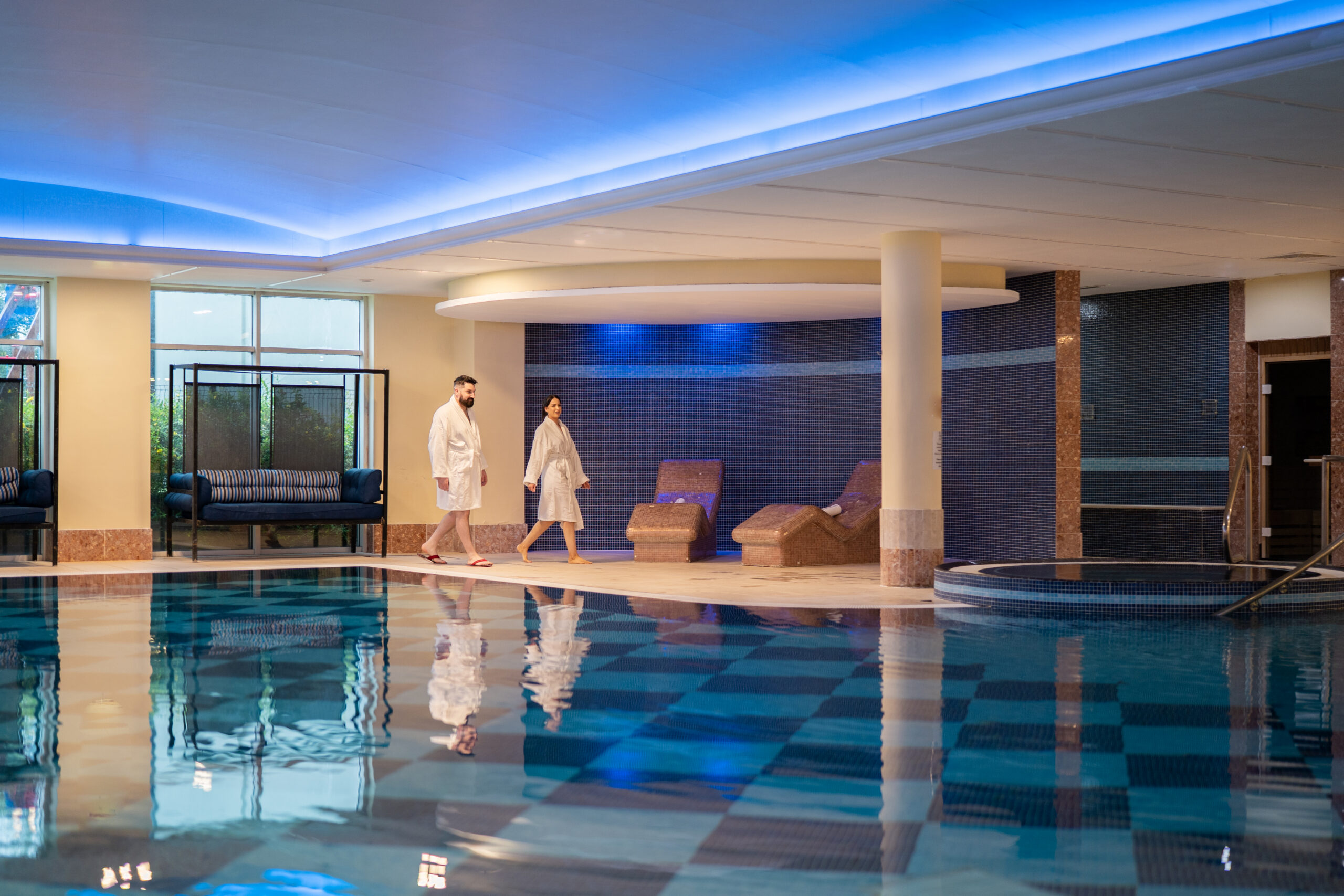 Two guests wearing bathrobes are walking towards the relaxation loungers beside the swimming pool at Hillgrove Hotel & spa Monaghan.